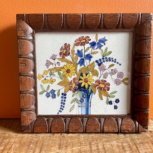 Könnte beinhalten: Eine gerahmte Fliese mit einem farbenfrohen Blumendesign. Die Fliese zeigt einen Blumenstrauß in einer blau-weißen Vase mit gelben, roten, blauen und weißen Blumen. Der Rahmen ist aus dunklem Holz mit einem geschnitzten Design.