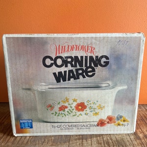 Puede incluir: Una caja blanca con un diseño floral en la parte delantera. La caja es para una cacerola cubierta Corning Ware de 1,5 litros. La caja tiene el texto "Wildflower Corning Ware" en la parte delantera. La caja también tiene el texto "1 1/2 qt. Covered Saucepan" y "A-116-7".