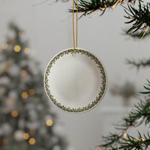 May include: A white ceramic Christmas ornament with a green leafy border. The ornament is round and has a hole at the top for hanging.