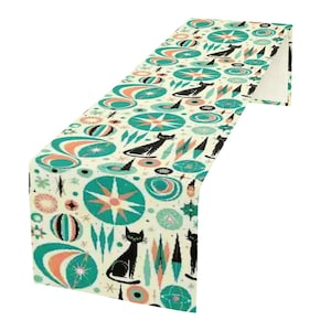 May include: A cream-colored table runner with a mid-century modern design. The pattern features black cat silhouettes, teal starbursts, and orange and teal atomic shapes. The runner is made of a soft fabric.