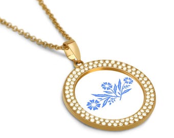 Puede incluir: Collar de oro con un colgante redondo que presenta un diseño floral azul sobre un fondo blanco. El colgante está rodeado de diamantes de imitación transparentes.