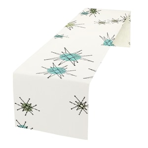 May include: White table runner featuring a repeating pattern of blue and green starbursts. The starbursts are outlined in black and have a retro mid-century modern design.