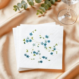 May include: A stack of white paper napkins with a delicate floral design featuring blue flowers and green leaves. The napkins are arranged on a beige satin surface, with a sprig of greenery and a wine glass in the background.