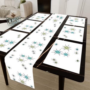 May include: A set of six white placemats and a table runner featuring a geometric star design in shades of blue and green. The placemats and runner are displayed on a black table.