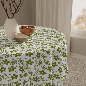 May include: A white round tablecloth with a green floral pattern. The tablecloth is on a round table with a bowl of fruit and a vase with a branch.