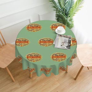 Op de afbeelding: Een ronde, lichtgroene tafelkleed met een herhalend patroon van vintage Fire-King Jade-ite logo's in rood en goud. Een witte theekop en een tijdschrift liggen op de tafel. Het tafelkleed ligt op een houten tafel.
