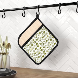 May include: A white pot holder with a green floral pattern. The pot holder has a black border and a beige backing. It is hanging on a black metal rack.