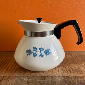 May include: A white vintage coffee pot with a black handle and lid. The pot features a silver band and a blue leaf design. The pot is sitting on a wooden surface with an orange background.