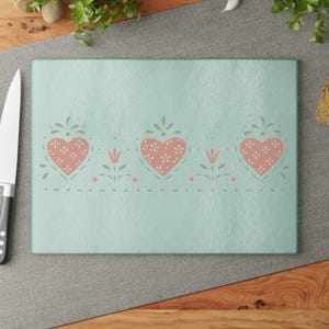 May include: A rectangular, pale turquoise chopping board featuring a decorative border of pink hearts filled with white floral patterns. The hearts are interspersed with small floral and dotted designs. A stainless steel knife is visible.