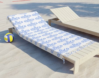 Op de afbeelding: Een witte strandhanddoek met een blauw bloemenpatroon en franjes. De handdoek ligt op een houten ligstoel op een zandstrand.