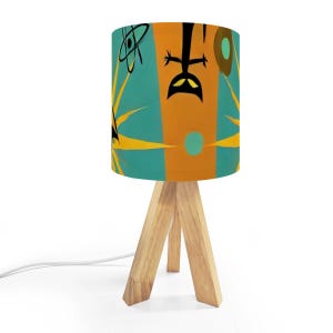 May include: A small table lamp with a cylindrical shade featuring a retro design in teal, orange, and yellow. The lamp has a wooden tripod base and a white power cord.