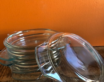 May include: A stack of clear glass Pyrex baking dishes with handles. The dishes are oval-shaped and appear to be vintage. The background is a solid orange color.