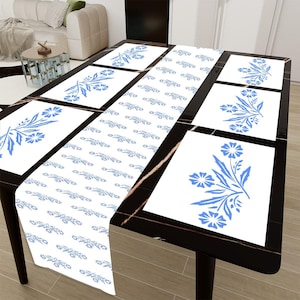 Puede incluir: Una mesa de comedor negra con un camino de mesa blanco y manteles individuales a juego. El camino de mesa y los manteles individuales presentan un diseño floral azul. La mesa está en una habitación bien iluminada.