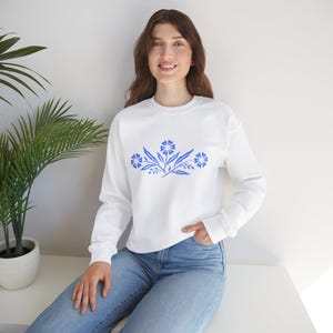 Könnte beinhalten: Weißer Pullover mit einem blauen Blumendesign auf der Vorderseite. Der Pullover hat lange Ärmel und einen Rundhalsausschnitt.