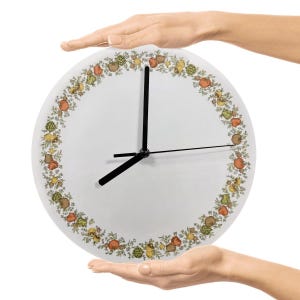 Könnte beinhalten: Weiße runde Uhr mit schwarzen Zeigern, gehalten von zwei Händen. Das Ziffernblatt hat einen Rand aus farbenfrohen Obst- und Gemüseillustrationen. Die Zeiger zeigen ungefähr 8:38 Uhr an.