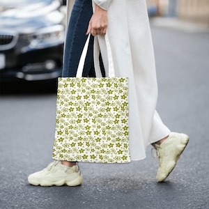 Op de afbeelding: Een witte tote bag met een groen bloemenpatroon. De tas heeft een witte handvat en wordt door een persoon gedragen.