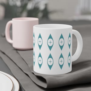 Può includere: Tazza in ceramica bianca con un motivo geometrico turchese di rombi con un segno più all'interno di ogni rombo. La tazza è appoggiata su una superficie grigia.