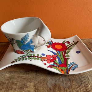 May include: A white ceramic teacup and saucer with a colorful floral and bird design. The cup has a unique curved handle and the saucer has a wavy edge.