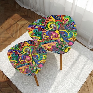 Op de afbeelding: Een set van twee kleurrijke, abstracte, bladvormige bijzettafels met houten poten. De tafelbladen hebben een psychedelisch patroon van swirls en lijnen in tinten roze, geel, blauw, groen en oranje.