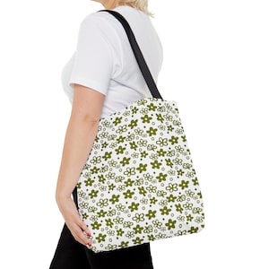 May include: A white tote bag with a green floral pattern. The bag has black straps and is being held by a person.