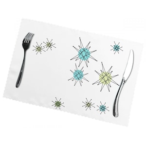 Op de afbeelding: Een wit placemat met een gegolfde rand met een herhalend patroon van zwarte en witte sterren met blauwe en groene accenten. Een zilveren vork en een botermes liggen op het placemat.