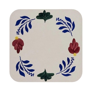 Puede incluir: Posavasos de cerámica blanca con un diseño floral azul, verde y rojo. El diseño presenta hojas y flores estilizadas alrededor del borde del posavasos.