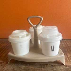 May include: A white plastic salt and pepper shaker set with a brown plastic holder. The shakers have the letters "P" and "M" printed on them.