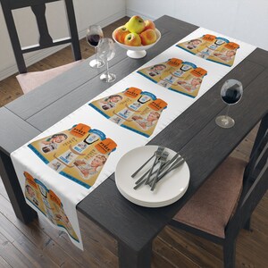 Può includere: Runner da tavolo bianco con un design pubblicitario vintage Pyrex. Il design include illustrazioni retrò di donne e pentole Pyrex, con il testo "Pyrex Flameware". Il runner è esposto su un tavolo di legno scuro.