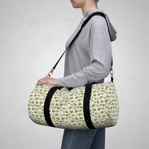 May include: A white and green duffel bag with a black strap and a floral pattern. The bag is being worn by a person.