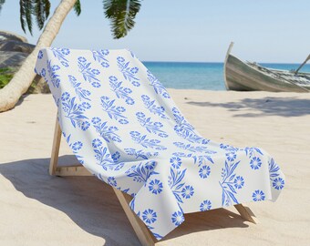 Op de afbeelding: Een witte strandstoel met een blauw bloemenpatroon op de stof. De stoel staat op een zandstrand met een houten boot op de achtergrond.