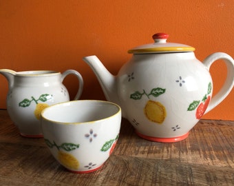 May include: A white teapot, creamer, and sugar bowl with a lemon and strawberry design. The teapot has a yellow lid and a red handle. The creamer and sugar bowl have red accents.