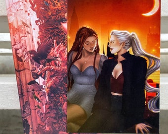 May include: Two art prints featuring illustrations of women. One print has a detailed, ornate design in shades of red and orange. The other print shows two women in a city setting under a crescent moon, with a warm, orange sky.