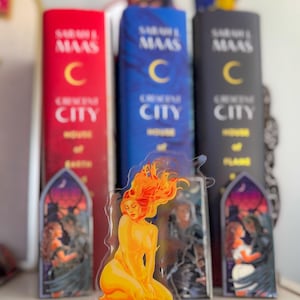 Peut inclure: Une figurine transparente représentant une femme aux cheveux orange flamboyants, agenouillée. Elle est placée devant trois livres intitulés "Crescent City" de Sarah J. Maas. Les livres sont rouges, bleus et noirs.