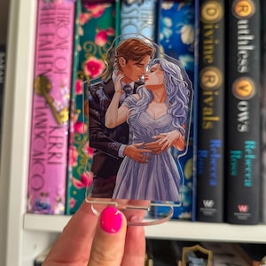 May include: A clear acrylic book stand with a romantic illustration of a kissing couple. The artwork shows a man in a suit and a woman in a gown. The stand is held in front of a bookshelf with book titles visible.