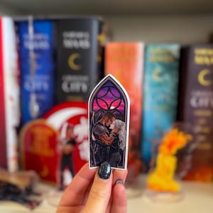 Peut inclure: Un autocollant représentant une illustration romantique d'un couple s'embrassant, sur fond de vitrail avec un coucher de soleil. L'autocollant a une forme d'arche gothique et est tenu devant un arrière-plan flou de livres.