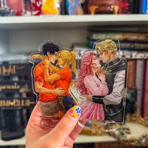 May include: Acrylic stand featuring two romantic illustrations. One shows a couple embracing, the other a couple gazing at each other. The figures have detailed features and are in vibrant colors. The stand is held in a hand with a floral nail design.