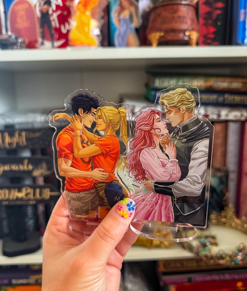 May include: Acrylic stand featuring two romantic illustrations. One shows a couple embracing, the other a couple gazing at each other. The figures have detailed features and are in vibrant colors. The stand is held in a hand with a floral nail design.