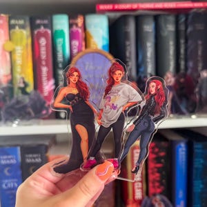 Puede incluir: Figuras acrílicas que representan tres ilustraciones de una mujer pelirroja. Una lleva un vestido negro, otra una camiseta y leggings, y la tercera una chaqueta de cuero y vaqueros. Las figuras se sostienen delante de una estantería.