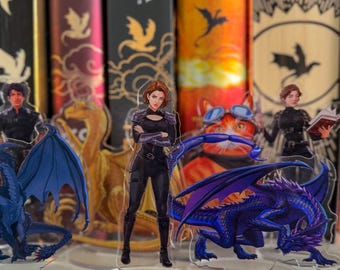 May include: Acrylic character standees featuring fantasy characters and dragons. The standees are in front of a shelf of books with the author's name "Rebecca Yarros" visible on the spines. The dragons are blue and purple.
