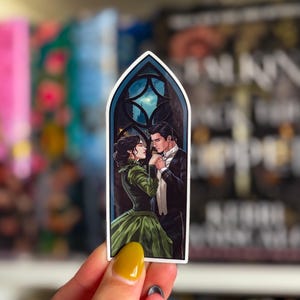 May include: A sticker depicting a romantic scene of a couple in formal wear, framed by a gothic window with a moonlit cityscape. The woman is in a green gown, and the man wears a tuxedo. The sticker has a white border.