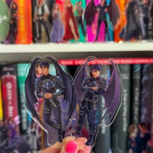 Może przedstawiać: Dwie akrylowe figurki postaci z ciemnymi włosami i dużymi fioletowymi skrzydłami. Postać ma na sobie ciemną zbroję i czarny strój. Figurki są trzymane przed półką z książkami i innymi figurkami.