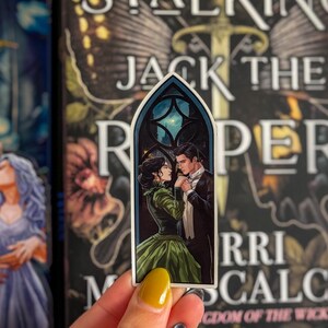 May include: A sticker depicting a romantic scene of a couple in Victorian dress, set against a night sky within a gothic window frame. The woman wears a green gown, and the man is in a tuxedo. The sticker is held in front of a book cover.