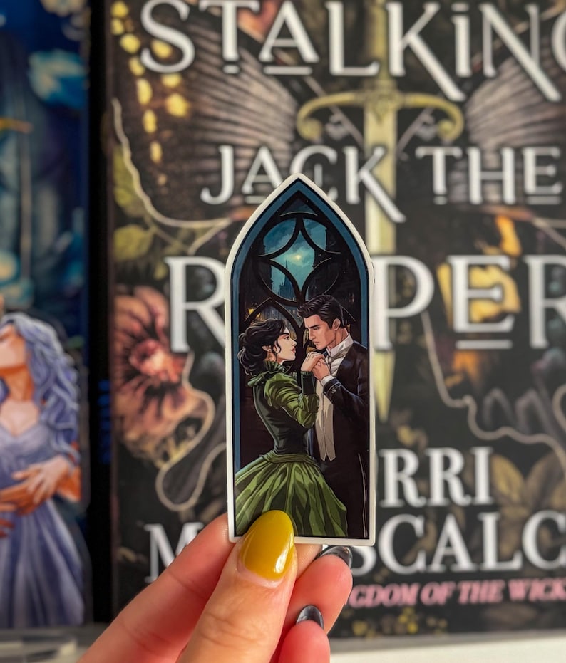 May include: A sticker depicting a romantic scene of a couple in Victorian dress, set against a night sky within a gothic window frame. The woman wears a green gown, and the man is in a tuxedo. The sticker is held in front of a book cover.