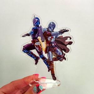 May include: A clear acrylic stand-up featuring two figures in Mandalorian armor. One figure is in blue and purple armor, the other in maroon and silver. Both are posed with weapons, set against a white background.