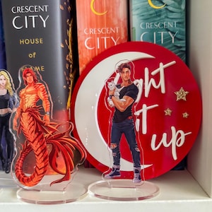 Puede incluir: Expositores acrílicos con personajes de la serie de libros "Crescent City". Uno representa a un personaje masculino con cola de tigre. Otro muestra a un personaje masculino sosteniendo un gato blanco, con las palabras "light it up" sobre un fondo rojo.