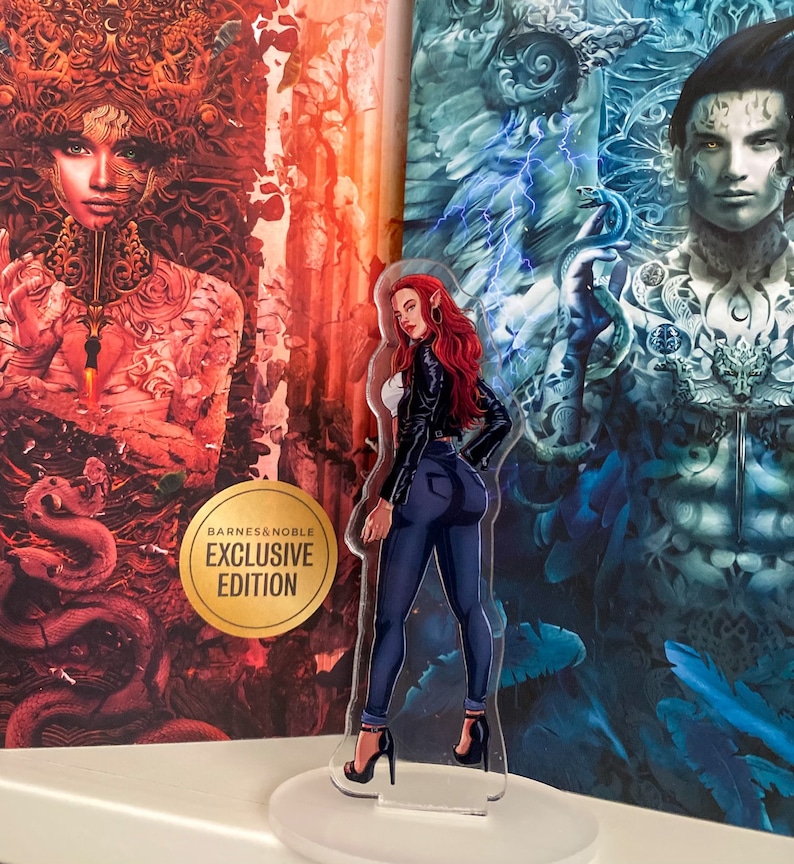 May include: An acrylic standee of a woman with red hair, wearing a black jacket, white top, and blue jeans, standing in front of two book covers. The standee is on a white base. The book covers have illustrations and the text "BARNES & NOBLE EXCLUSIVE EDITION".