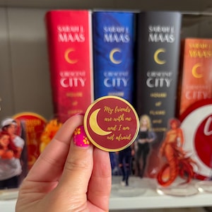 Peut inclure: Un badge rond bordeaux avec une bordure dorée et un croissant de lune. Le badge porte l'inscription "My friends are with me and I am not afraid." Des livres sont visibles en arrière-plan.