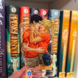 May include: A clear acrylic charm featuring a couple embracing, both wearing orange shirts. The charm has a blue baseball cap with a white logo. The background shows a bookshelf with several books.