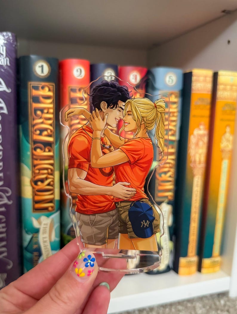 May include: A clear acrylic charm featuring a couple embracing, both wearing orange shirts. The charm has a blue baseball cap with a white logo. The background shows a bookshelf with several books.