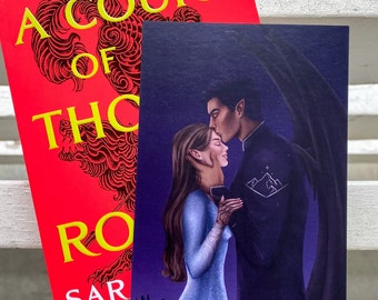 May include: A red book titled "A Court of Thorns and Roses" by Sarah J. Maas, with a matching art print. The print features a couple embracing, with a dark-haired man kissing a woman's forehead. The book cover has a dragon illustration.